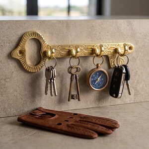 May include: A gold-toned metal key-shaped wall hook rack featuring an ornate engraved pattern. Four hooks hold a set of silver keys, an antique-style skeleton key, a round blue-faced compass, and a black car key fob. A brown leather glove lies below.