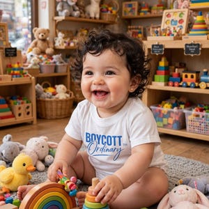 May include: A white short-sleeved bodysuit with the blue text Boycott Ordinary printed on the front. The garment is worn by a smiling child sitting on a gray rug surrounded by various wooden toys, including a rainbow stacker and blocks.