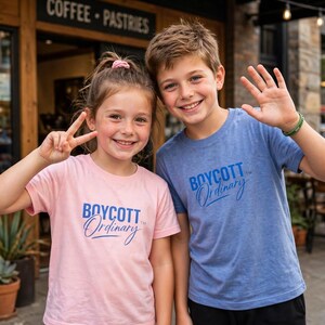 May include: A light pink t-shirt and a heather blue t-shirt, both featuring the text BOYCOTT Ordinary in blue lettering. The shirts have short sleeves and a crew neckline, made of a soft fabric, and are worn by two children smiling at the camera.