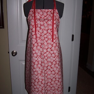 May include: A red and white patterned apron with a peppermint swirl design. The apron has red straps and trim. The apron is displayed on a mannequin.