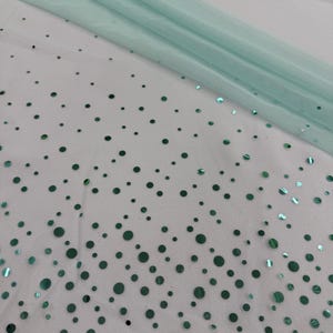 May include: A close-up of a white fabric with scattered, iridescent teal-green sequins. A sheer, light teal fabric is draped across the top. The sequins vary in size and are scattered across the fabric, creating a shimmering effect.