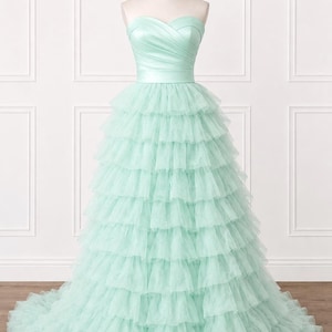 May include: A floor-length strapless gown in a soft mint green color. The bodice features a sweetheart neckline with diagonal pleated fabric. The skirt is constructed from multiple horizontal layers of ruffled tulle, creating a full silhouette.