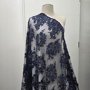 May include: Navy blue lace fabric with floral embroidery and beaded accents. The delicate lace is draped over a mannequin, showcasing the intricate details of the design. The fabric is ideal for creating elegant apparel.