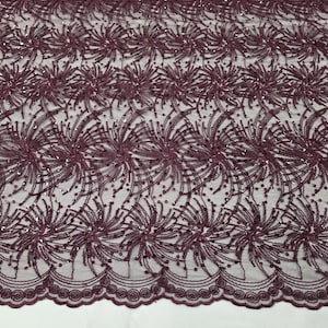 May include: Burgundy lace fabric with a floral design. The fabric features a repeating pattern of stylized flowers and scattered sequins. The bottom edge has a scalloped trim. The fabric is semi-transparent.