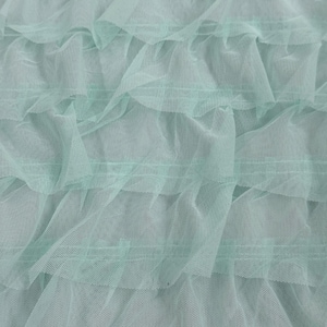 May include: A light mint green fabric featuring multiple horizontal layers of sheer, fine-mesh ruffles. The material is translucent and gathered to create a textured, wavy appearance across the entire surface of the textile.
