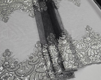 Könnte beinhalten: Eine Rolle schwarzes Tüllgewebe mit silberner, paillettenbesetzter floraler Spitzenborte. Die Spitze weist komplizierte Designs auf und ist stark mit schimmernden Pailletten verziert. Der Stoff ist gefaltet und zeigt die Textur und Details.