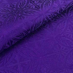 May include: Close-up of a deep purple fabric with a repeating floral and geometric pattern. The fabric has a textured surface and a slight sheen, with folds adding depth to the design. The pattern is darker purple.