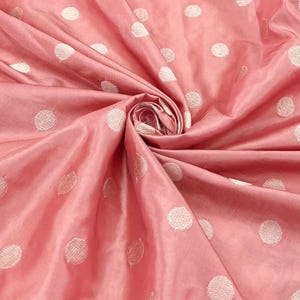 May include: A close-up of a folded, pink fabric with a satin sheen. The fabric is adorned with large, white, embroidered polka dots. The fabric is gathered in a spiral, showcasing its texture and color.