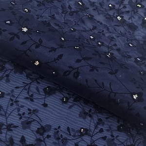 May include: Navy blue sheer fabric with a floral pattern. The fabric features dark floral embroidery and scattered silver sequins. The material is folded, showing the texture and depth of the design. This textile is suitable for apparel and craft projects.