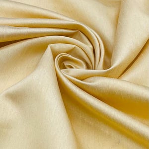 May include: A length of solid light gold fabric with a subtle sheen and a fine, horizontal slubbed texture. The material is gathered into a central swirl, creating soft, flowing folds and deep shadows that highlight the smooth, lustrous surface.
