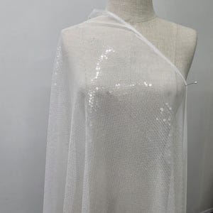 May include: White sequin fabric draped over a mannequin. The fabric is sheer, with small, round sequins evenly spaced across its surface. The sequins catch the light, creating a shimmering effect. The fabric is pinned to the mannequin.