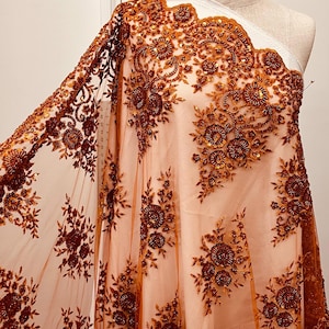 May include: A sheer, peach-colored fabric with intricate floral embroidery in shades of brown and copper. The design features detailed beaded flowers and leaves, creating a textured, elegant look. The fabric is draped over a mannequin, showcasing its flowing quality.