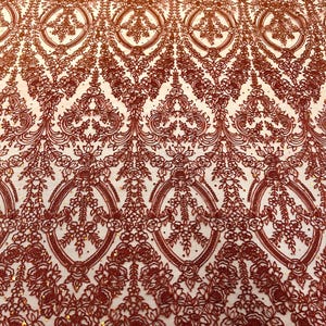 May include: Close-up of a detailed, reddish-brown lace fabric. The intricate design features floral and geometric patterns, including oval shapes and delicate flower arrangements. The lace has a scalloped edge and a subtle shimmer, creating a luxurious texture.