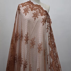 May include: Brown lace fabric with a floral design and a dotted background. The lace is draped over a mannequin, showcasing the intricate details and texture of the material. The fabric has a scalloped edge.