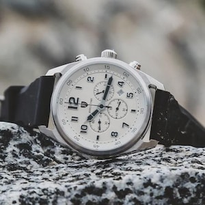 May include: A silver-toned wristwatch with a white face and black hands and numerals. The watch has a black band and multiple sub-dials. The watch is sitting on a textured, gray surface.