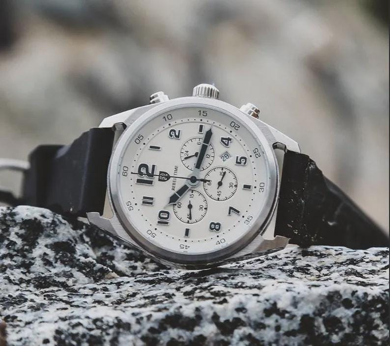 May include: A silver-toned wristwatch with a white face and black hands and numerals. The watch has a black band and multiple sub-dials. The watch is sitting on a textured, gray surface.