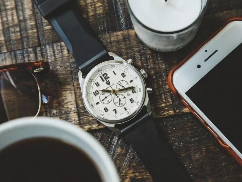 May include: A silver wristwatch with a white face and black band rests on a wooden surface. The watch has multiple dials and black numerals. Other items include a smartphone, sunglasses, a candle, and a cup of coffee.