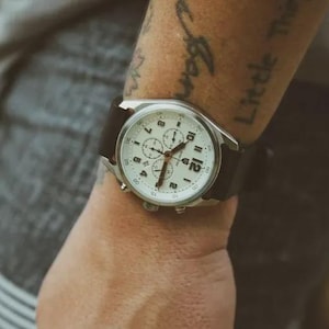 May include: A silver-toned wristwatch with a white face and black leather band. The watch features multiple subdials and a date display. The watch is on a wrist.