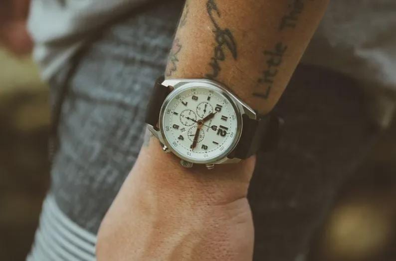 May include: A silver-toned wristwatch with a white face and black leather band. The watch features multiple subdials and a date display. The watch is on a wrist.