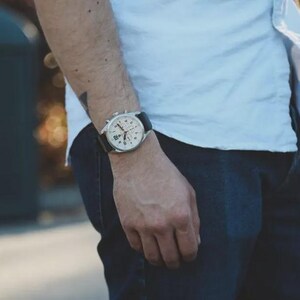 May include: A silver-toned chronograph watch with a black leather band is displayed on a person's wrist. The watch features a white face with black numerals and sub-dials. The person is wearing a white shirt and dark blue jeans.