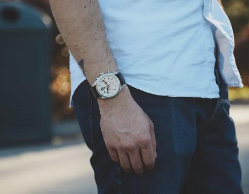 May include: A silver-toned chronograph watch with a black leather band is displayed on a person's wrist. The watch features a white face with black numerals and sub-dials. The person is wearing a white shirt and dark blue jeans.