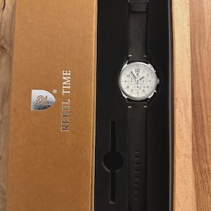 May include: A wristwatch with a white face and black leather band is displayed in a brown box. The watch has silver-colored accents and a buckle. The box is labeled "REBEL TIME" and has a logo.