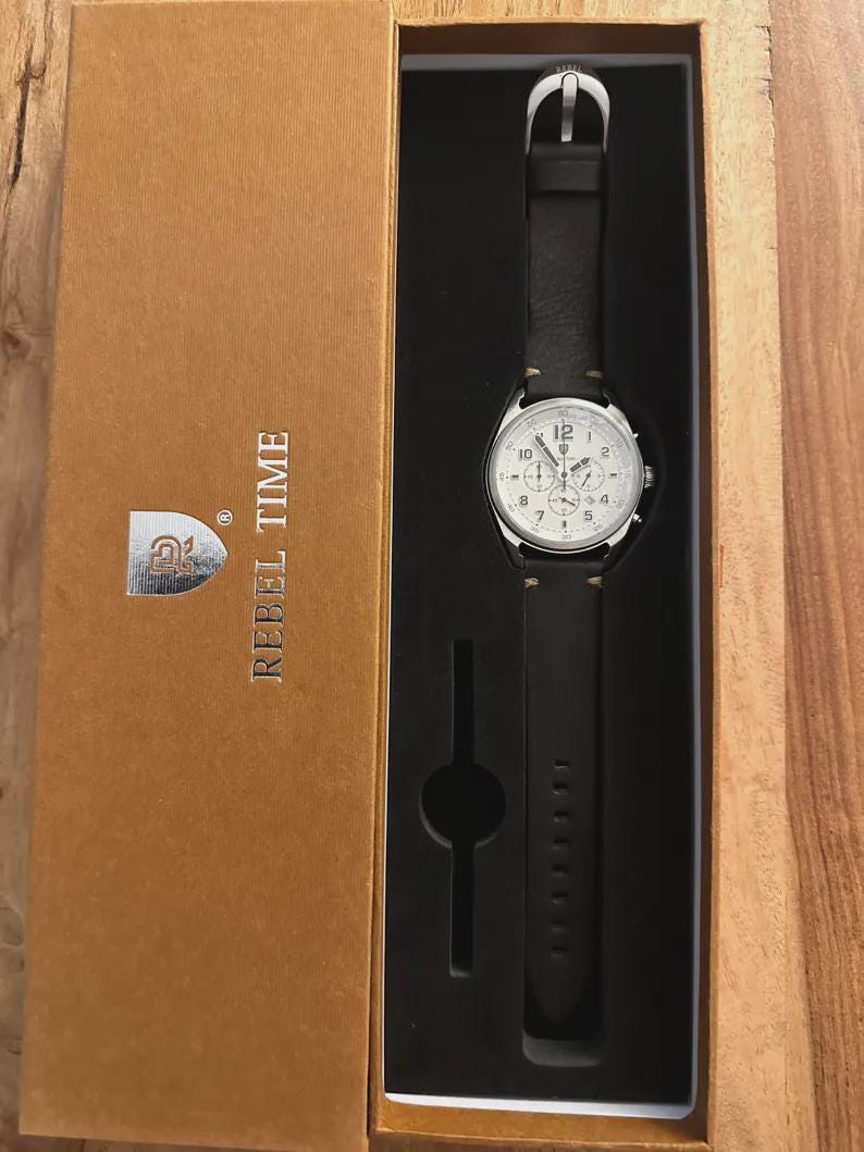 May include: A wristwatch with a white face and black leather band is displayed in a brown box. The watch has silver-colored accents and a buckle. The box is labeled "REBEL TIME" and has a logo.