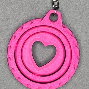 May include: A pink keychain with a heart-shaped cutout in the center. The keychain has a textured, circular design.
