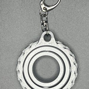May include: A silver-colored metal keychain with a circular design. The circle has two concentric rings with a decorative pattern around the outer edge.