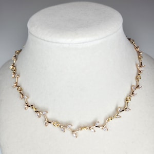 May include: A gold-toned necklace with a delicate vine design, adorned with small, clear gemstones. The necklace is displayed on a white mannequin bust, showcasing its elegant and nature-inspired aesthetic. The necklace is a perfect accessory for special occasions.