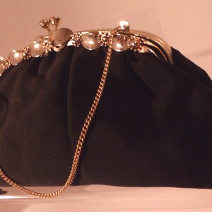 May include: A black clutch purse with a gold-colored chain strap. The purse has a decorative clasp with clear, gem-like accents. The top edge of the purse is also adorned with similar gems.
