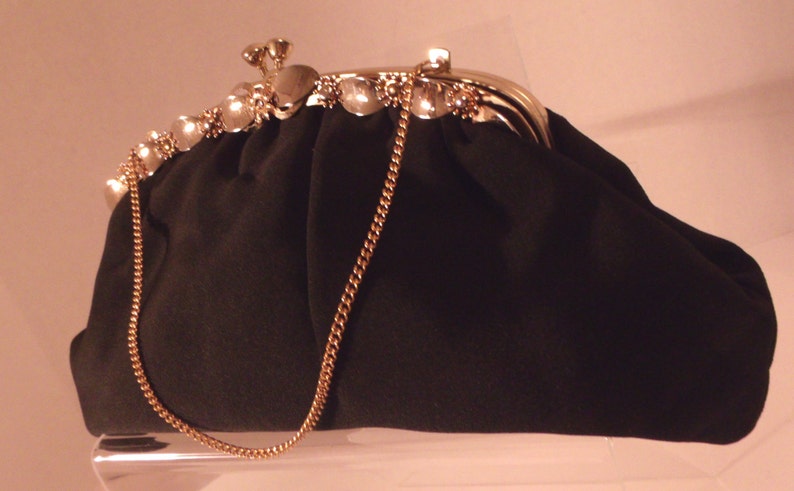 May include: A black clutch purse with a gold-colored chain strap. The purse has a decorative clasp with clear, gem-like accents. The top edge of the purse is also adorned with similar gems.