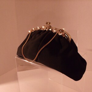May include: Black clutch purse with a gold chain strap. The clasp is adorned with pearls and a decorative gold element. The purse is displayed on a clear acrylic stand.