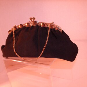 May include: A black vintage handbag with a gold-colored clasp and chain strap. The clasp is adorned with small, round gold accents. The bag is made of a textured fabric and sits on a clear display stand.