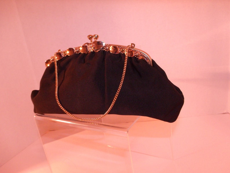 May include: A black vintage handbag with a gold-colored clasp and chain strap. The clasp is adorned with small, round gold accents. The bag is made of a textured fabric and sits on a clear display stand.