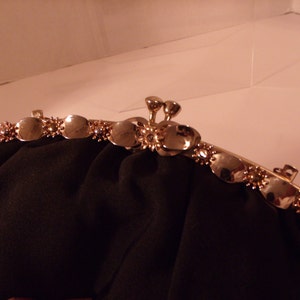 May include: A black evening bag with a gold-toned clasp. The clasp features a series of decorative, oval-shaped accents with small, sparkling embellishments. The bag's fabric is a dark, matte material.