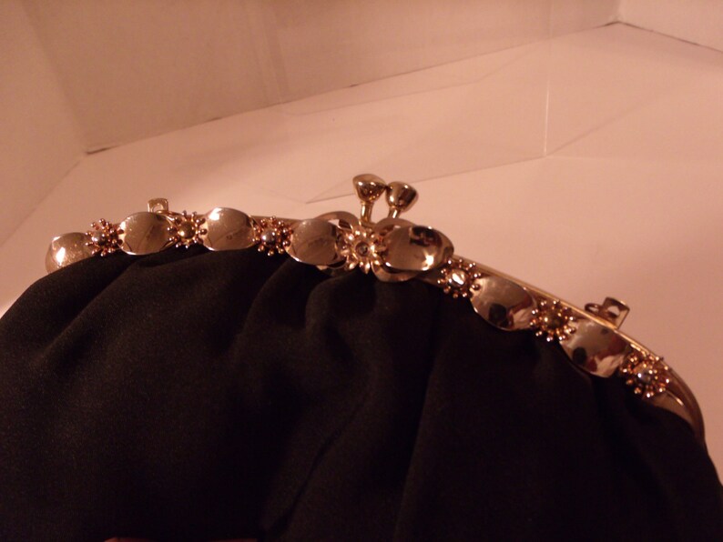 May include: A black evening bag with a gold-toned clasp. The clasp features a series of decorative, oval-shaped accents with small, sparkling embellishments. The bag's fabric is a dark, matte material.