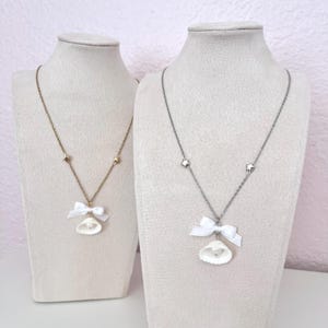 以下が含まれることがあります： マネキンのバストに飾られた2つのネックレス。1つは星のアクセントが付いたゴールドチェーンと、白いリボンが付いた貝殻のペンダント。もう1つは星のアクセントが付いたシルバーチェーンと、白いリボンが付いた貝殻のペンダント。