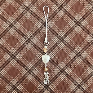 May include: A silver charm with a white heart pendant, small orange beads, and a deer charm. The charm is attached to a white cord with a silver loop. The background is a brown plaid pattern.