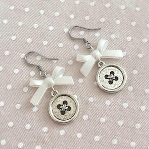 May include: A pair of button earrings with a white bow and a silver-colored button charm. The button charm has a cross-shaped design in the center. The earrings hang from silver-colored fish hook ear wires. The background is a light pink fabric with white polka dots.