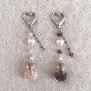 May include: Two heart-shaped keychains with silver clasps. Each keychain features a small glass bottle filled with gemstones, a heart-shaped charm, and a chain of small hearts. One bottle contains pink stones, the other contains dark red stones.