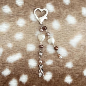 May include: A silver-toned heart-shaped lobster clasp keychain featuring two dangling chains. One chain includes brown and cream beads, a textured heart charm, and a small deer charm. The second chain features brown and cream beads and a smooth heart charm.