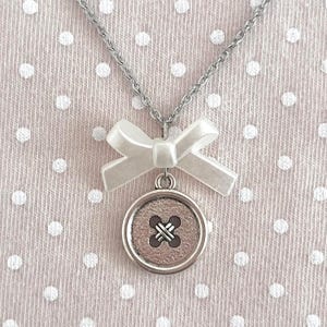 May include: A silver-toned necklace featuring a button pendant with a cross-stitch design. A white bow is attached above the button. The necklace hangs on a silver chain against a pink background with white polka dots.