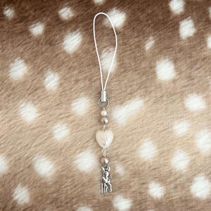 Puede incluir: Un colgante para teléfono con un lazo de cordón blanco, una pequeña anilla de metal plateado, una cuenta blanca nacarada, una cuenta de color topo metalizado, una cuenta en forma de corazón color crema con relieve y un dije de metal plateado de un cervatillo.