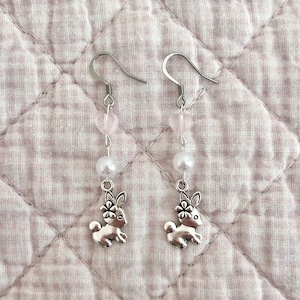 May include: A pair of silver dangle earrings featuring a small bunny charm, a white pearl bead, and a pink heart-shaped bead.