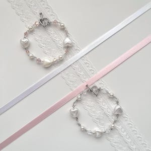 Könnte beinhalten: Zwei Armbänder mit herzförmigen Charms, Perlen und Kristallperlen. Die Armbänder haben silberne Verschlüsse und liegen auf weißer Spitze mit weißen und rosa Bändern.