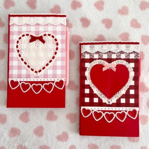 Peut inclure: Deux cahiers rouges avec des motifs en forme de cœur. L'un a un motif vichy rose, l'autre un motif vichy rouge. Les deux présentent de la dentelle blanche, des cœurs et un stylo rose.