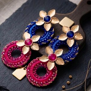 Puede incluir: Un par de pendientes colgantes con aros circulares envueltos en cuentas de color azul y rosa vibrantes. Cada aro está acentuado con una flor de metal dorado con una piedra preciosa central azul o rosa. Un pequeño dije cuadrado dorado cuelga.
