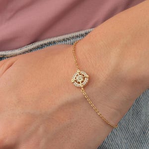 May include: A gold-colored bracelet featuring a compass rose design. The compass rose is encrusted with small, clear gemstones. The bracelet is delicate and has a thin chain. The person is wearing a pink shirt and blue jeans.