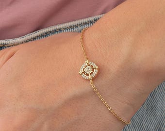 May include: A gold-colored bracelet featuring a compass rose design. The compass rose is encrusted with small, clear gemstones. The bracelet is delicate and has a thin chain. The person is wearing a pink shirt and blue jeans.